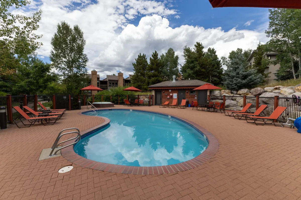 This image showcases an expansive outdoor swimming pool surrounded by comfortable lounge chairs, umbrellas, and lush greenery, with a backdrop of natural scenery. The setting invites guests to relax, swim, or socialize in a sun-soaked, resort-style environment.