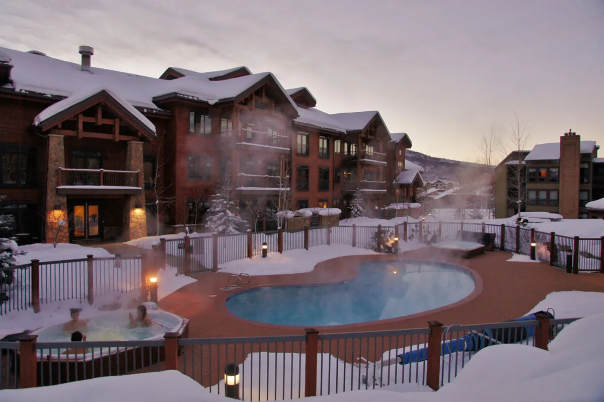 This image showcases a heated outdoor pool and hot tub surrounded by snow, with majestic mountain views and the building warmly lit in the background. Social spaces like a fire pit are visible, inviting gatherings and relaxation after a day on the slopes.