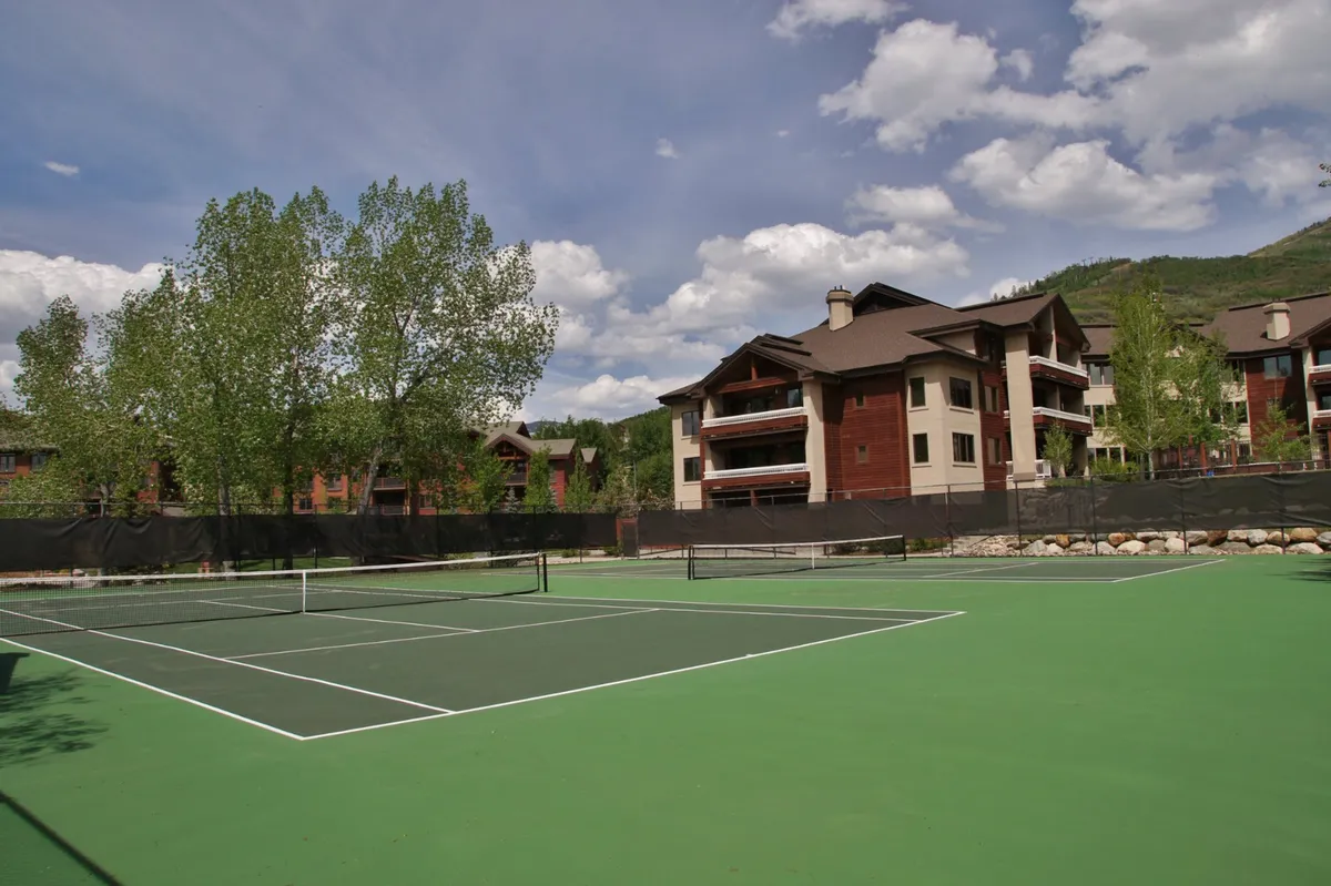 A vibrant view of tennis courts framed by tall green trees and a dramatic mountain backdrop. Residential buildings hint at proximity to accommodations, while people play tennis and enjoy the sunshine, capturing outdoor recreation and social energy.