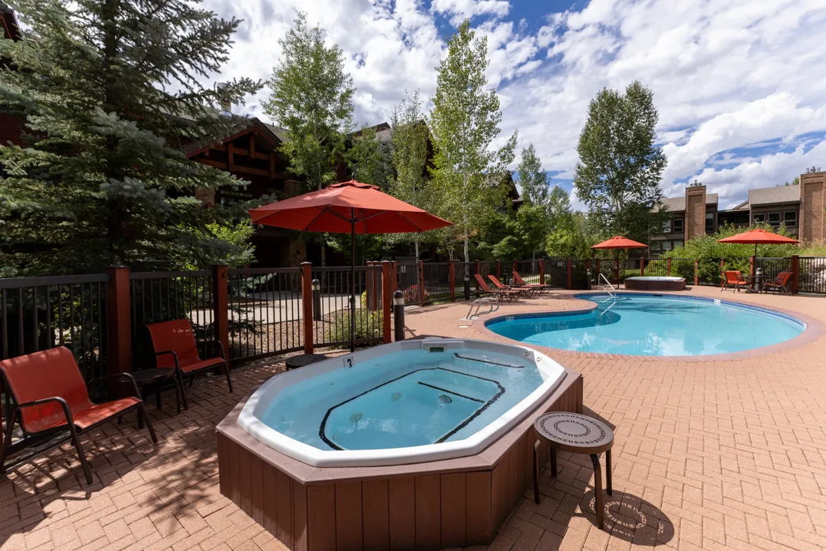 This exterior image features a heated outdoor swimming pool and hot tub surrounded by red umbrellas, lounge chairs, and lush trees. The tranquil water and cozy seating areas invite relaxation while showcasing the resort's leisure amenities against a scenic natural backdrop.