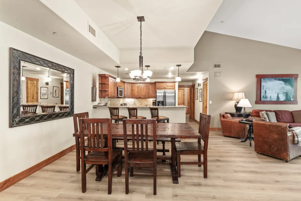 A welcoming open-concept interior view featuring a spacious dining table, a modern kitchen area, and plush living room seating. The scene is anchored by a mirror reflecting the warm ambiance, and the arrangement encourages social gatherings and shared meals. The overall mood is cozy and inviting, with visible connections between the main gathering spaces.