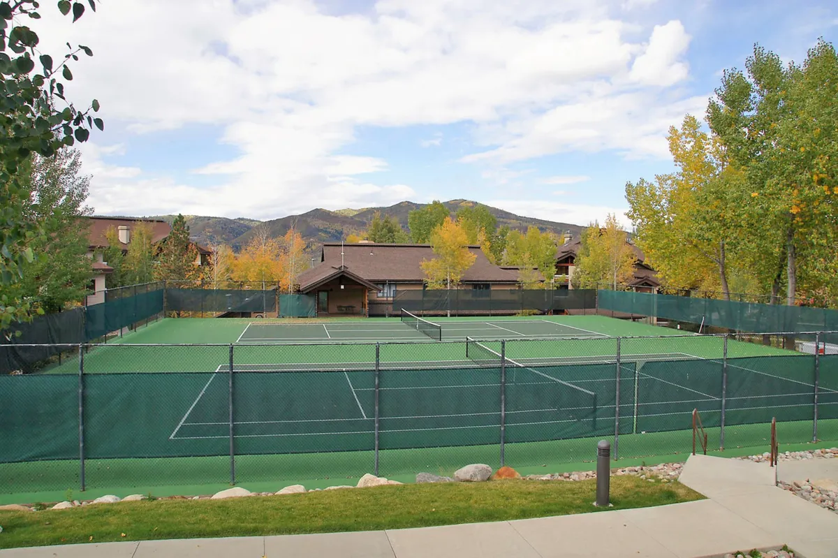 A well-maintained set of tennis courts framed by trees and with scenic views of the mountains beyond. The adjacent building and natural surroundings evoke a sense of community and outdoor recreation.