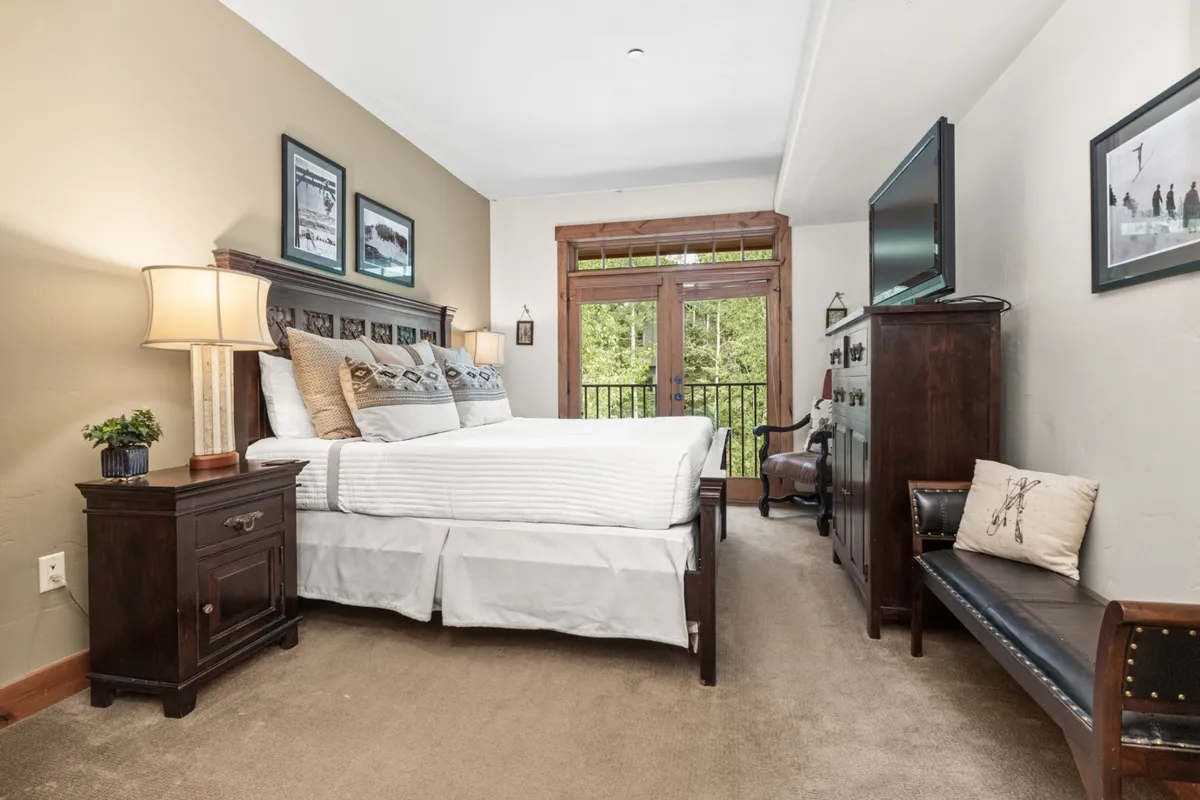 A tranquil bedroom with a plush bed, balcony doors that flood the space with daylight, nightstands, and framed pictures. The room is decorated for comfort and relaxation, with a serene and inviting mood and a TV visible for private viewing.