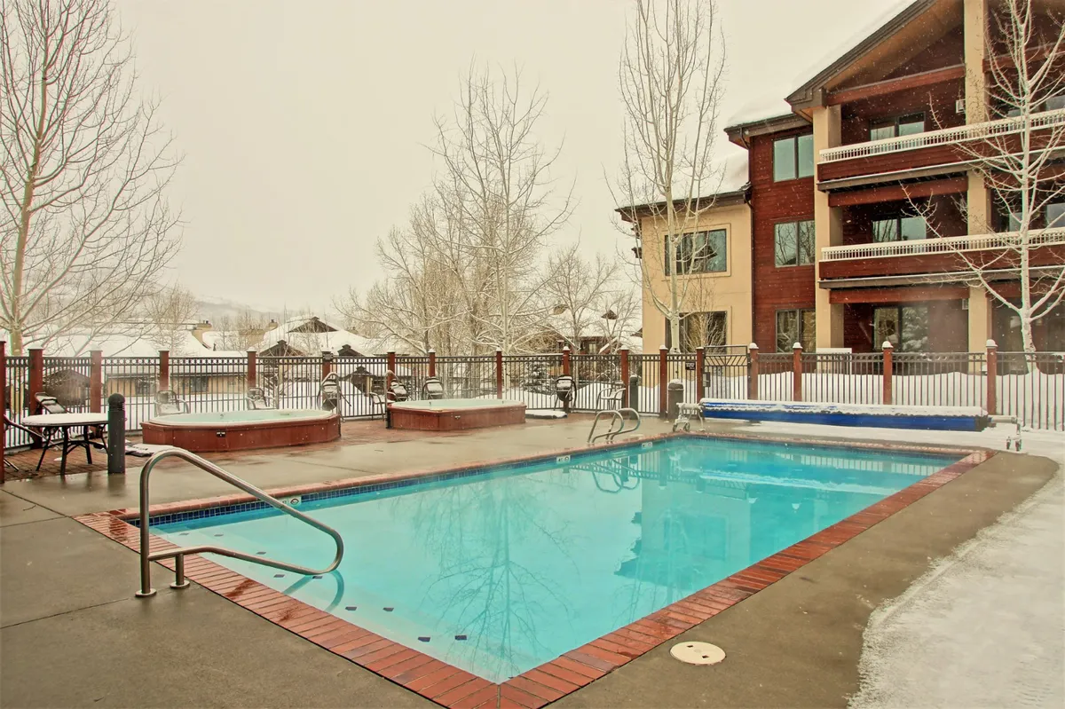 A stunning winter view of the resort's outdoor swimming pool and multiple hot tubs, surrounded by snow-covered grounds and framed by the building's facade. The steam rising from the hot tubs and the inviting glow of lights evoke warmth and relaxation even in the coldest months.