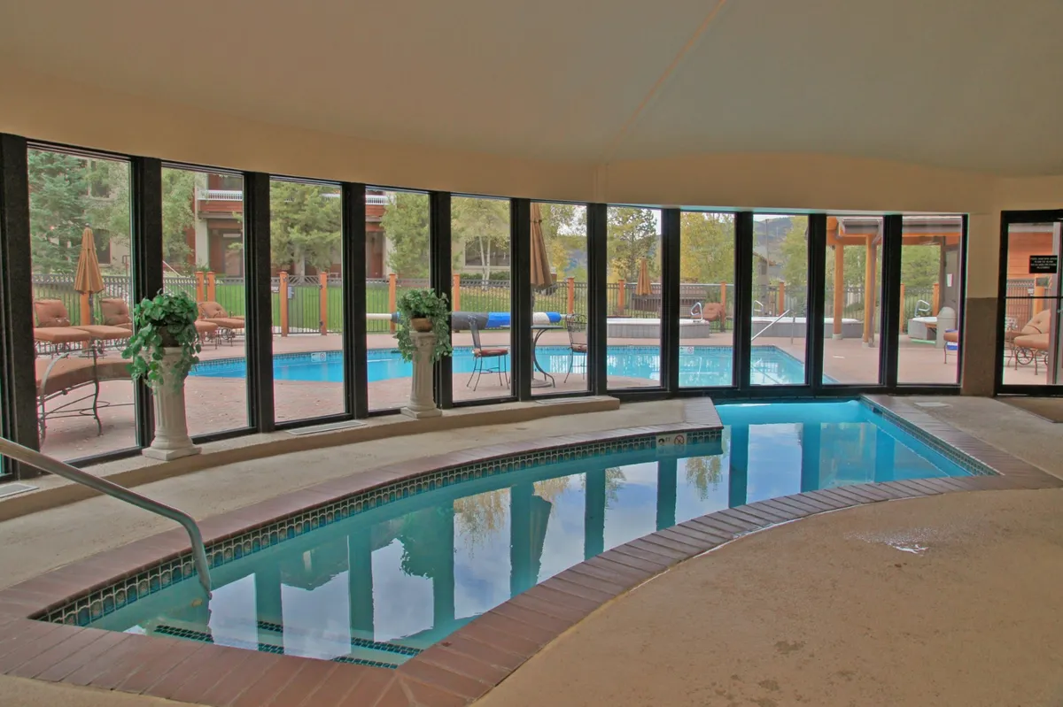 An inviting indoor pool area with a jacuzzi, large windows letting in natural light, lush potted plants, and views of the outdoor pool beyond. The tranquil setting is perfect for unwinding after a day of mountain adventures.