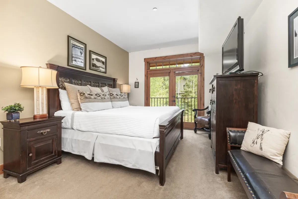 A spacious bedroom featuring a king-sized bed with upscale linens, a wooden nightstand, balcony doors letting in abundant natural light, and a decorative armchair. The space feels cozy, serene, and designed for both relaxation and comfort. There is a TV positioned for viewing from the bed, and the room opens out to a private balcony with views.