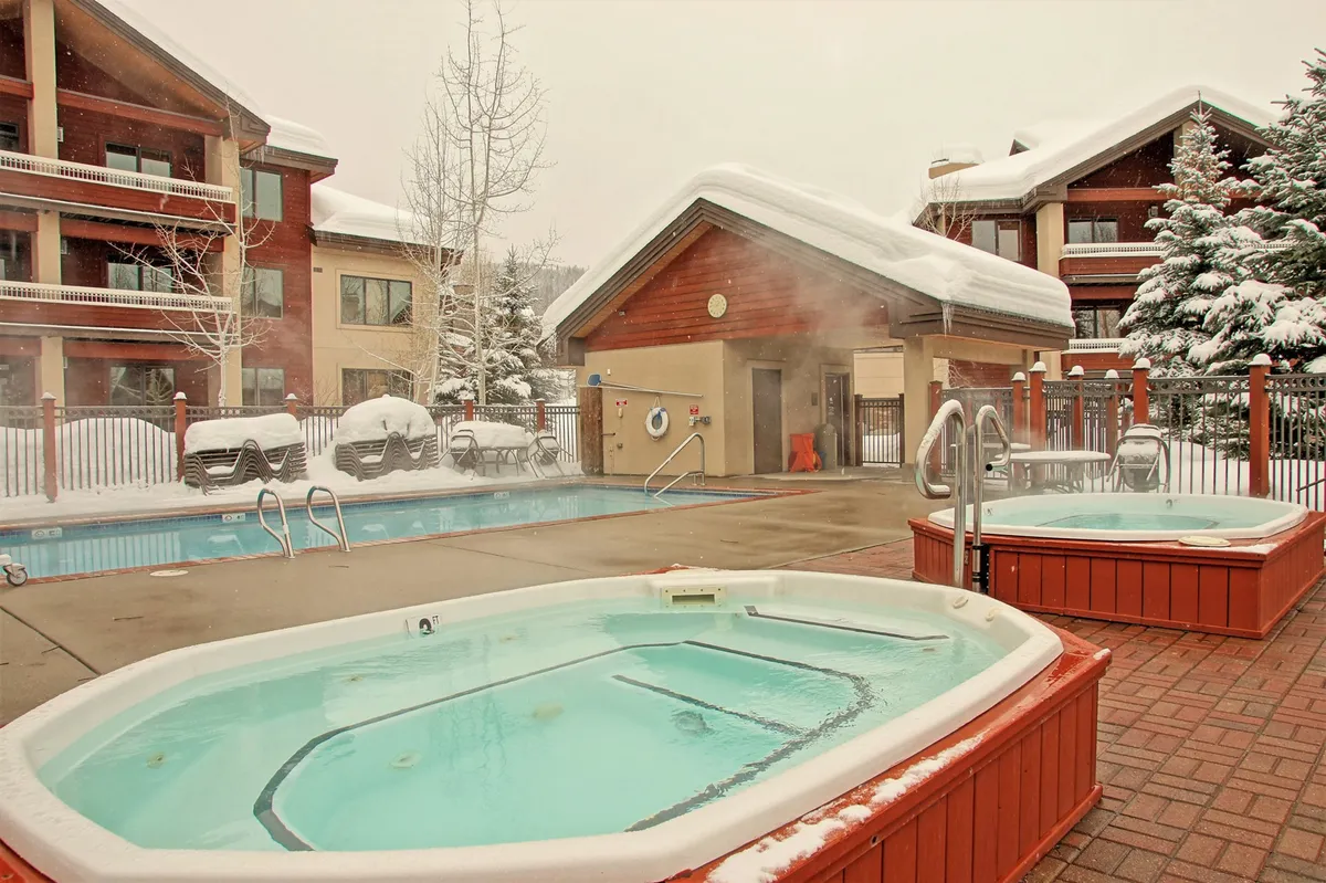 A snow-dusted outdoor pool area features two steaming hot tubs and a large swimming pool, with a building facade in the background and serene winter scenery surrounding the space. Friends and families can be seen enjoying the warm waters, contrasting the crisp mountain air.