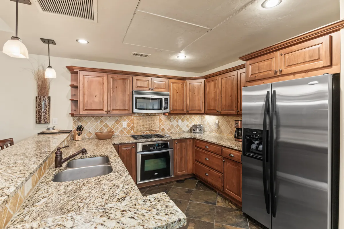 A modern kitchen with a granite countertop, wood cabinetry, stainless steel appliances, and stylish pendant lighting. The space is designed for both cooking and gathering, with a peninsula bar ideal for après-ski snacks or casual conversation. The warm, welcoming mood matches the section's depiction of a chef-inspired kitchen.